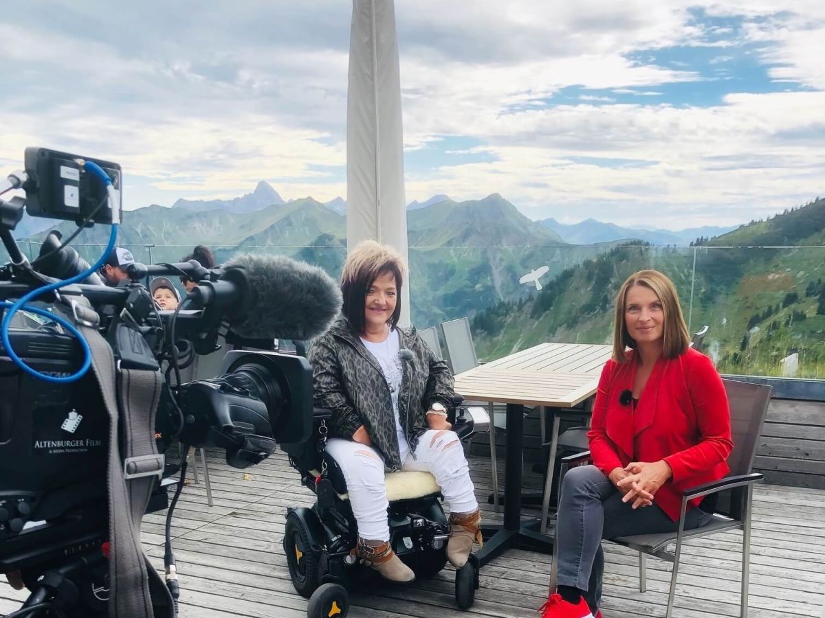 Marianne Hengl und Barbara Stöckl auf einer Terasse. Im Vordergrund eine Kamera, im Hintergrund eine Berglandschaft.